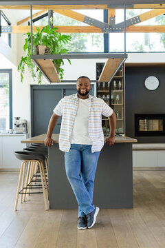 African man standing and leaning on central kitchen island, smiling near coffee machine and plants