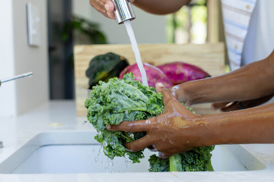 Curly kale is being rinsed under chrome faucet over white sink in kitchen, wooden crate behind