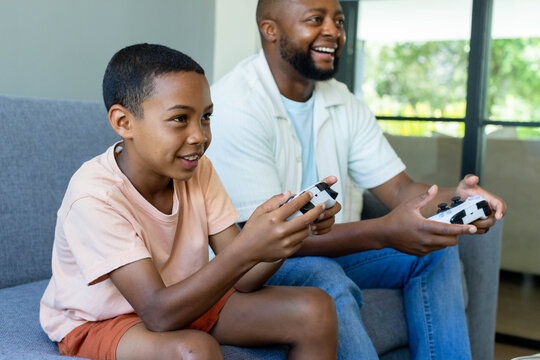 African American father and teenage son playing console games at home holding white controllers