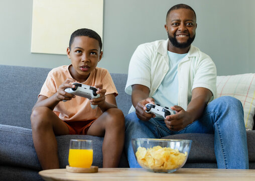 African American father-son sitting on couch at home holding white pads, chips, OJ, in casual wear
