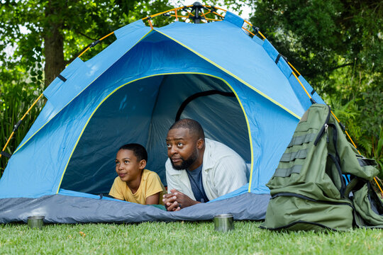 African American father and son lying, peering at blue tent opening on lawn with backpack, cups