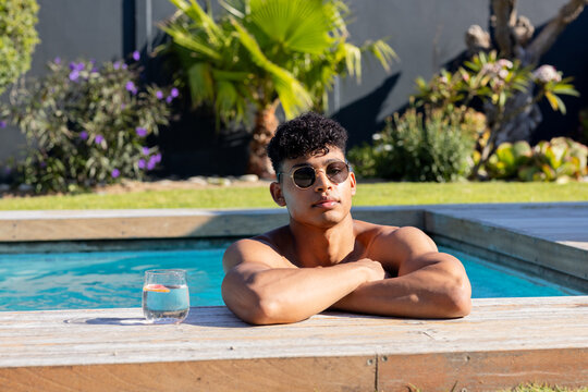 African American man leaning on backyard pool deck edge, wearing sunglasses, citrus glass to left