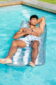 African American man shirtless striped trunks lounging on float in home pool holding glass