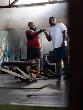 Two African American men fist-bumping while training in gym near weight bench, wearing shorts