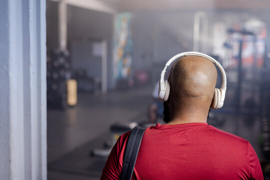 Man standing wearing red T-shirt and white headphones, carrying bag strap near weight plates in gym