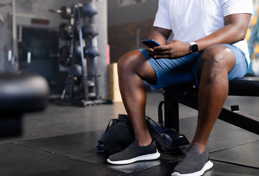 Man sitting on flat weight bench in gym training area, holding smartphone and wearing smartwatch