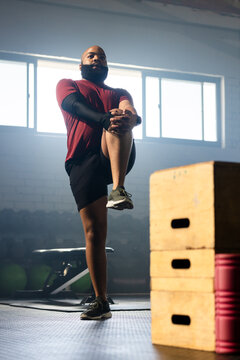 African American man wearing maroon shirt balancing knee-hugging in gym with plyo boxes foam roller
