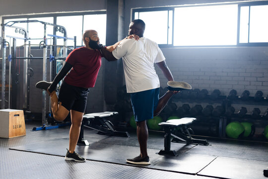 African male friends stretching quads on mats at gym, maroon and white shirts supporting each other