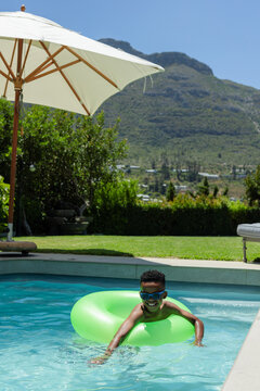 Male child floating on green inflatable ring in backyard pool under white umbrella wearing goggles