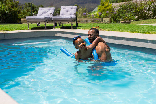 African American father and youth son playing and laughing in sunlit inground pool with blue noodle