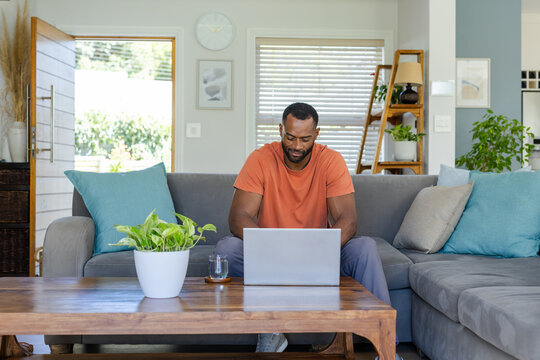African American man sitting in living room on gray sofa using silver laptop on coffee table