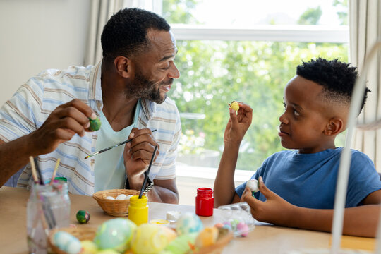 African American male father and son painting eggs at wooden table by window with brushes