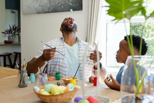 African American father and son painting and tossing decorated eggs at kitchen table with brushes