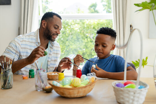 African American father and youth son painting eggs at home wooden table, guiding with brushes