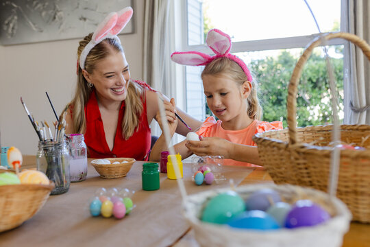 Mom and daughter wearing bunny-ear headbands painting eggs at craft table by window with paint jars