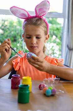 Child 7 sitting at table by window wearing orange-top pink-bunny-headband painting egg with brush