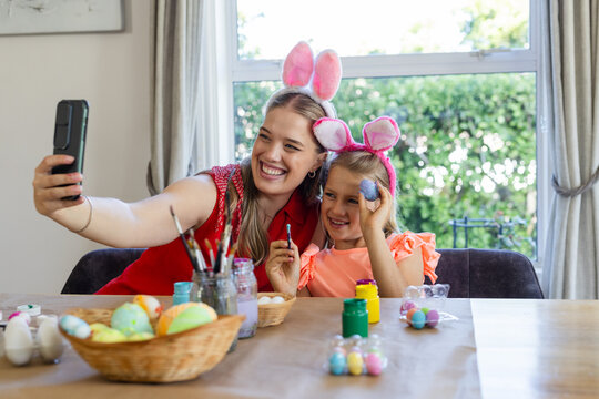 Mother in red top, daughter wearing pink bunny headbands painting eggs at home taking phone selfie