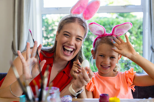 Mother and daughter wearing bunny-ear headbands, painting white egg with brushes at table by window