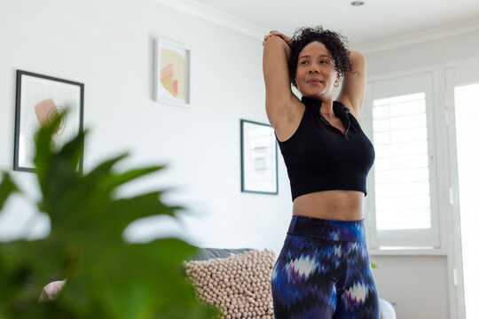 African American woman stretching arms overhead in living room wearing sportswear by couch cushion