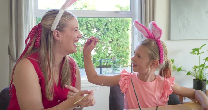 Mother and daughter painting eggs at window table, child reaching and painting mom's nose playfully