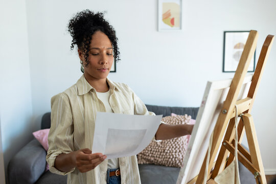 African woman standing holding reference and studying canvas on easel in living room, wearing jeans