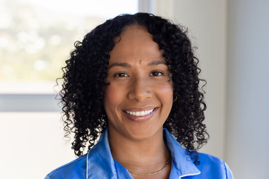 African adult woman smiling, looking at camera in bright room with window wearing blue satin shirt