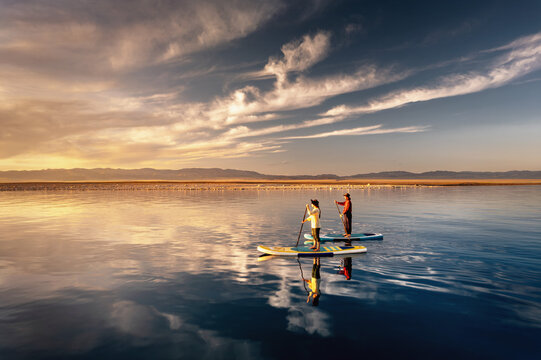 Two women paddleboarding on calm mountain lake at sunset, enjoying peaceful summer travel and freedom. Aerial view with reflection.