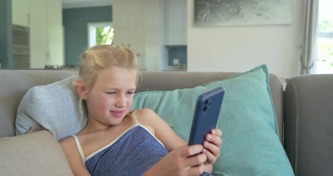 Preteen girl reclining on sofa wearing blue top, noticing phone screen, smiling and framing selfie
