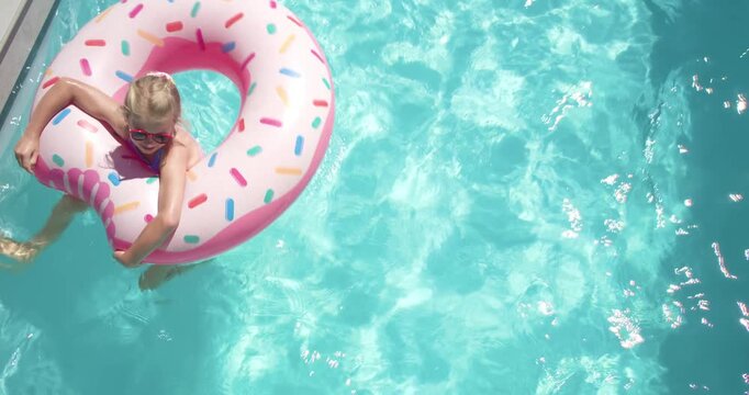 Sunlight causing child girl in swimsuit paddling pink donut float wearing sunglasses in pool