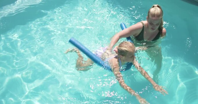 Adult woman placing blue noodle holding child girl practicing front float and kicks in home pool