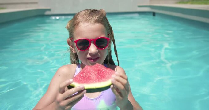 Girl in pastel swimsuit and red shades in backyard pool inspecting watermelon and biting to cool