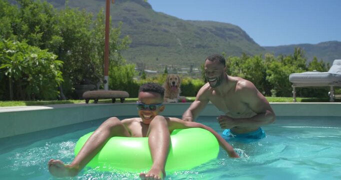 African American father in blue trunks steadying son, goggles on, guiding ring into backyard pool