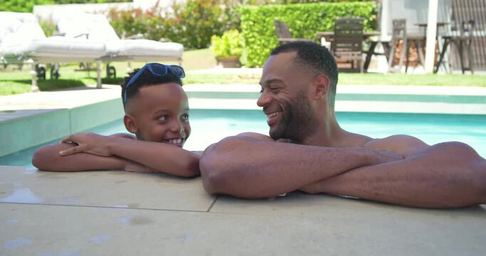 Father and teenage son leaning on backyard pool, meeting eyes, bonding in swimwear with sunglasses