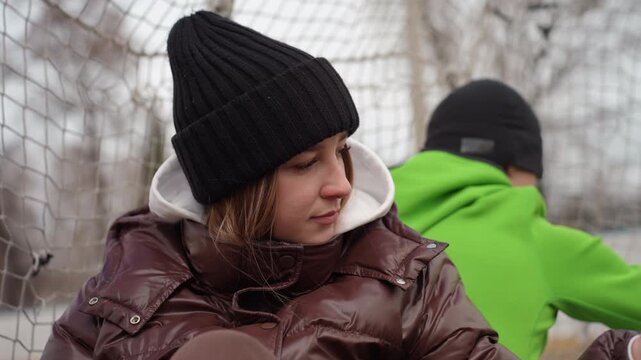 teen athlete thoughtful, teen girl in winter jacket watching teammate during practice break, youthful female athlete in beanie reflecting after practice near sports gear and sidelines