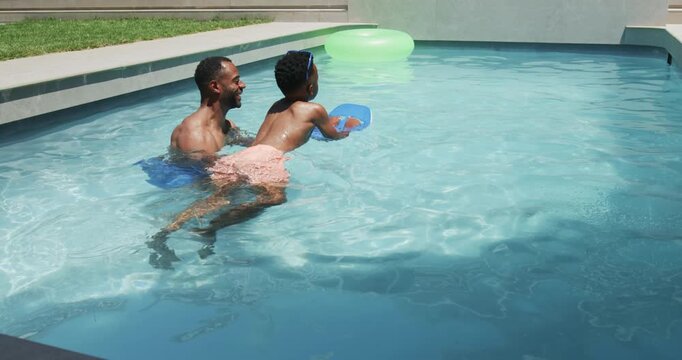 African father and son practicing kicking with blue kickboard in backyard pool toward lime ring