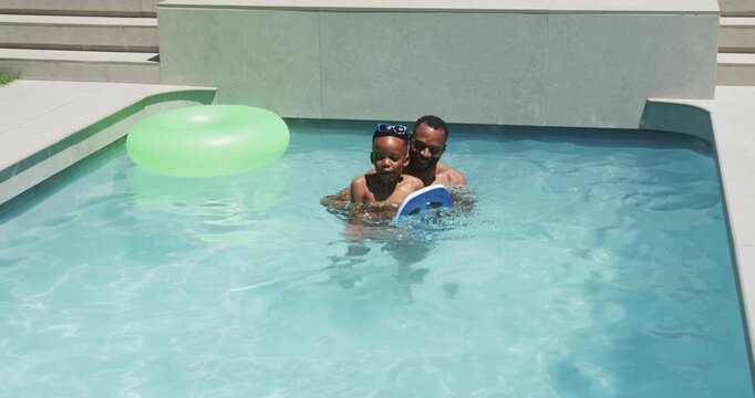 Adult is holding child in pool, guiding kicking practice with blue kickboard near green ring