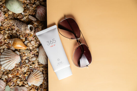 Top view of a white sunscreen tube labeled "365 Tone-Up" with aviator sunglasses on a beige background and sea shells on sand. Summer vacation and skin protection concept.