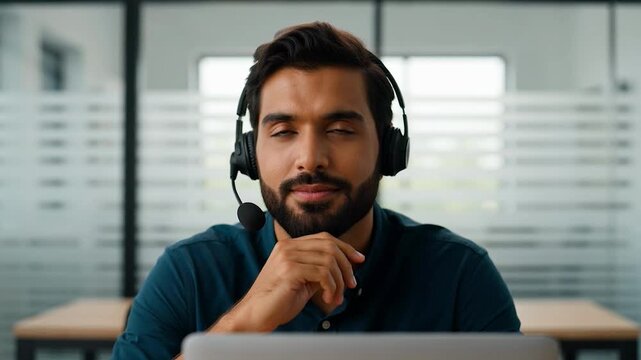Smiling man wearing a headset sitting at a desk in an office environment with a laptop