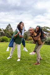 Fototapeta premium African American family laughing on garden lawn, parents giving piggyback rides with sneakers