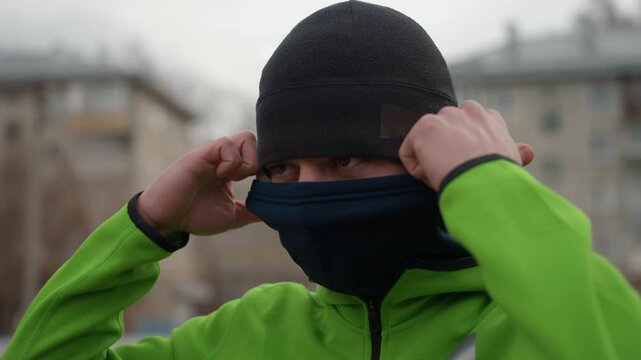 caucasian man tightening balaclava on city street green jacket beanie apartment blocks gray sky hands adjusting fabric determined expression chilly breath preparing for outdoor run