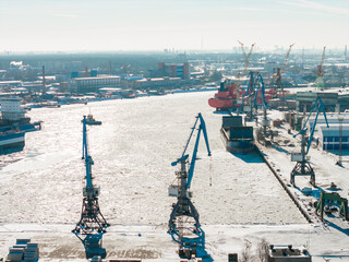 Aerial view shows Riga Shipyard in Riga, Latvia, with blue harbor cranes, icy quays, cargo vessels, and drifting ice, as warehouses and smokestacks fade in hazy sun. © Aerial Film Studio