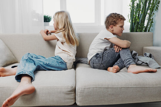 Two children sitting back to back on sofa looking upset and avoiding communication in bright living room. Concept of sibling conflict and family tension.