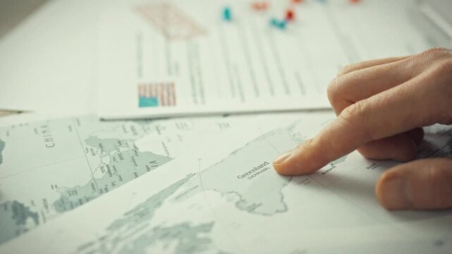 Planner enthusiastically gestures towards Greenland on sprawling world atlas, surrounded by documents vibrant navigation pins, utilizing various symbols markings to track coordinates on expansive map