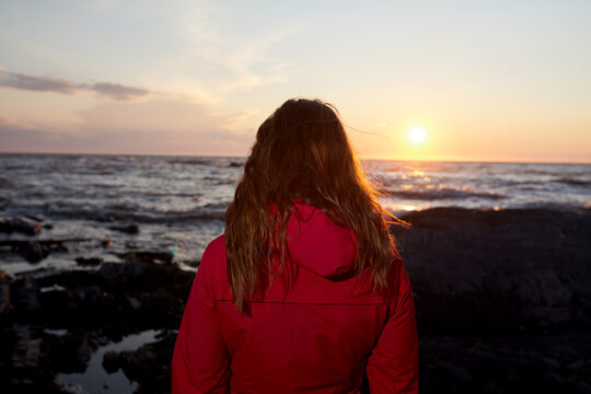 Woman in Red Jacket Standing on Rocky Shore at Sunset
