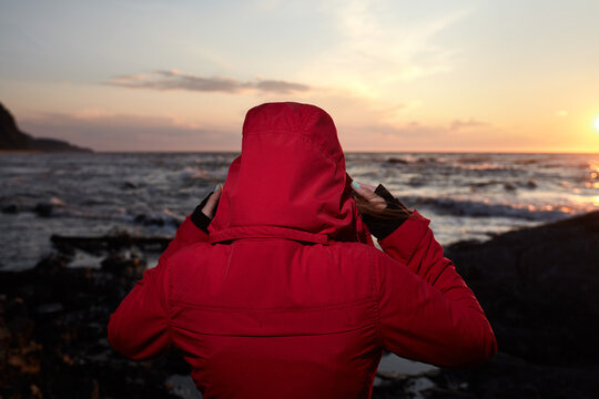 Woman in Red Jacket Standing on Rocky Shore at Sunset