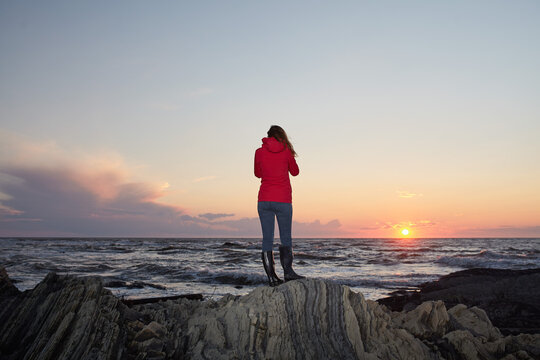 Woman in Red Jacket Standing on Rocky Shore at Sunset