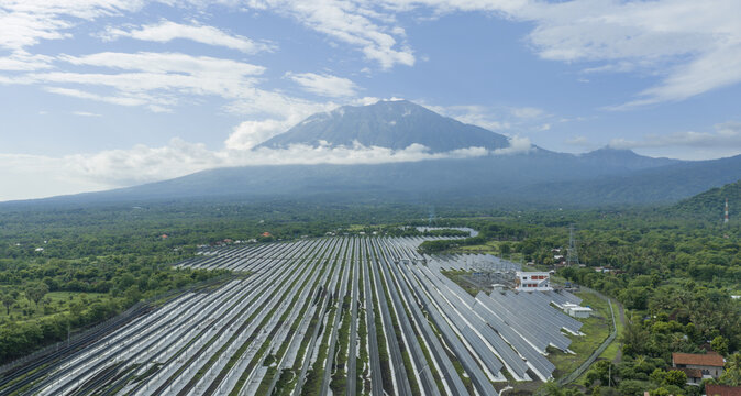 Aerial view of a vast solar farm contrasting with the lush green landscape and the majestic Mount Agung shrouded in clouds, Kubu, Bali, Indonesia.