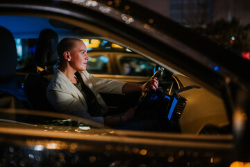 Woman driving car in urban setting at night. City lights illuminating the street and reflecting on the window