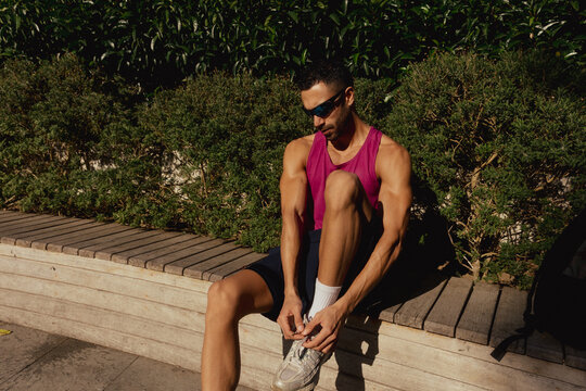 A young man is sitting on a park bench, tying his sneakers.