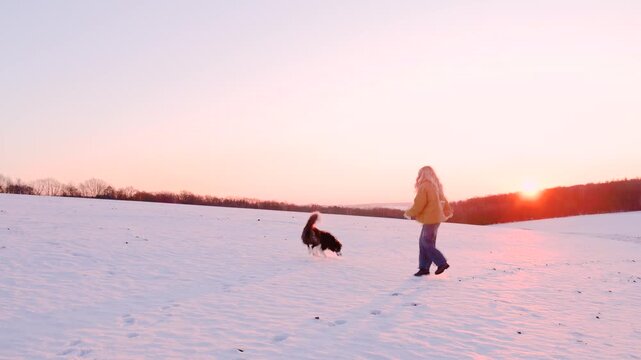 Young woman with long hair playing with her pet border collie dog in the snow, having fun at sunset with happy smile
